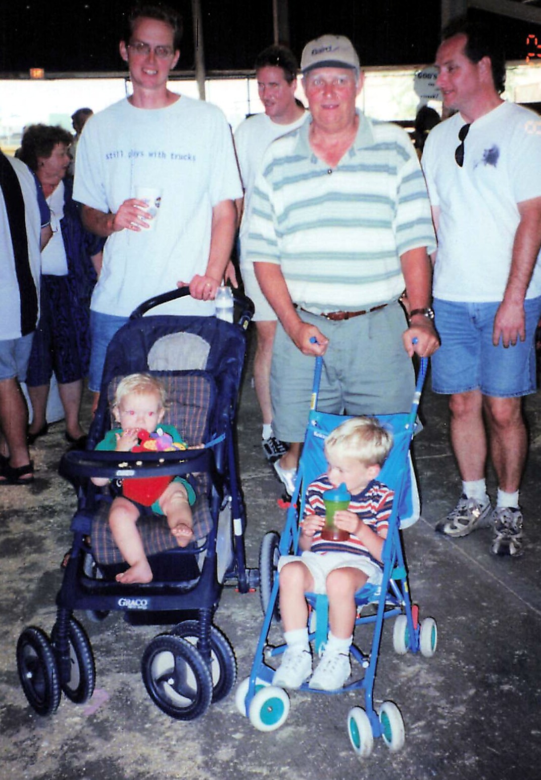 Group of adults standing behind two toddlers in pushchairs at a nighttime outdoor event.