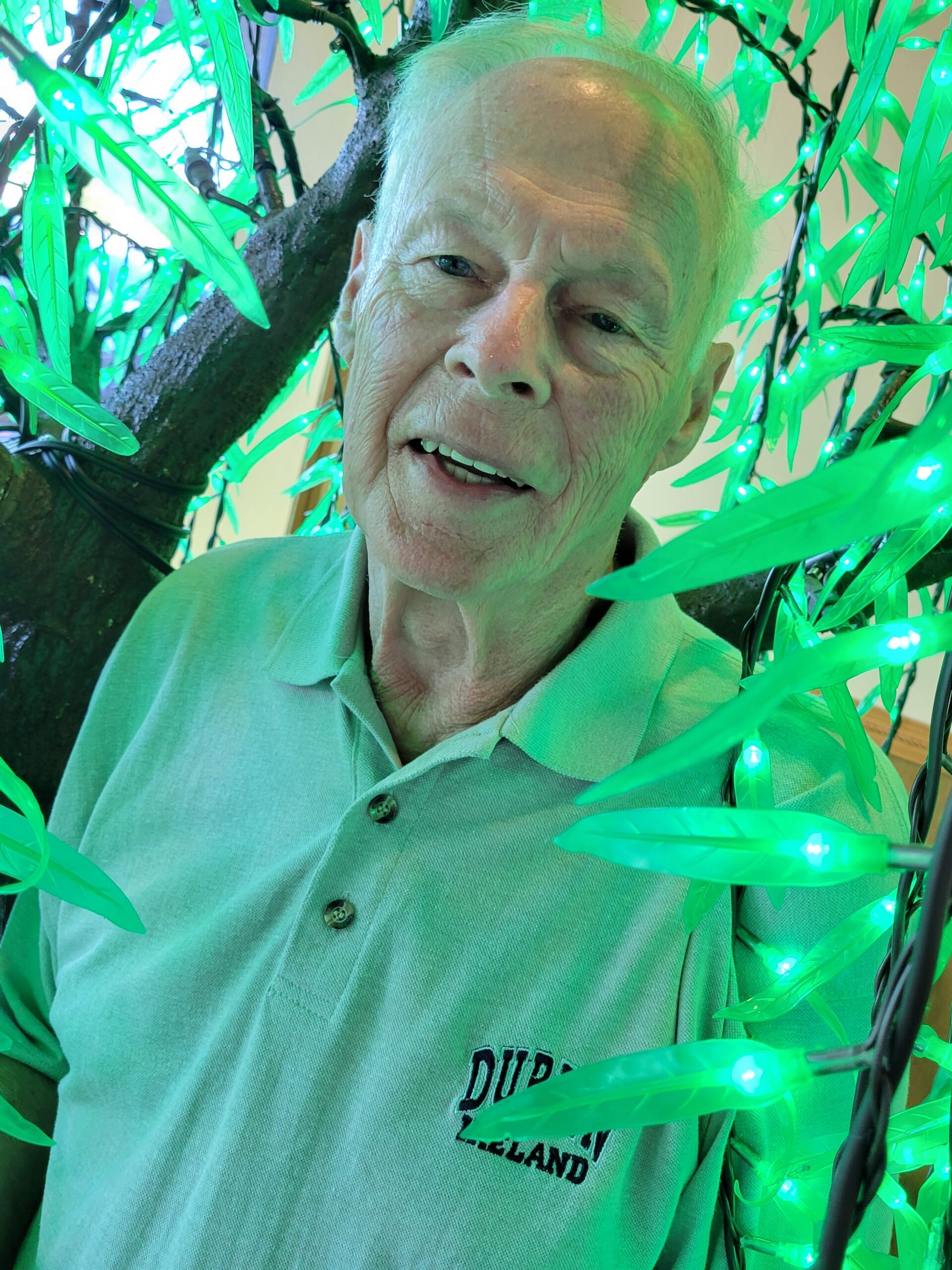 Close-up of an elderly man with short white hair, smiling, among glow-green string lights wrapped around a tree branch.