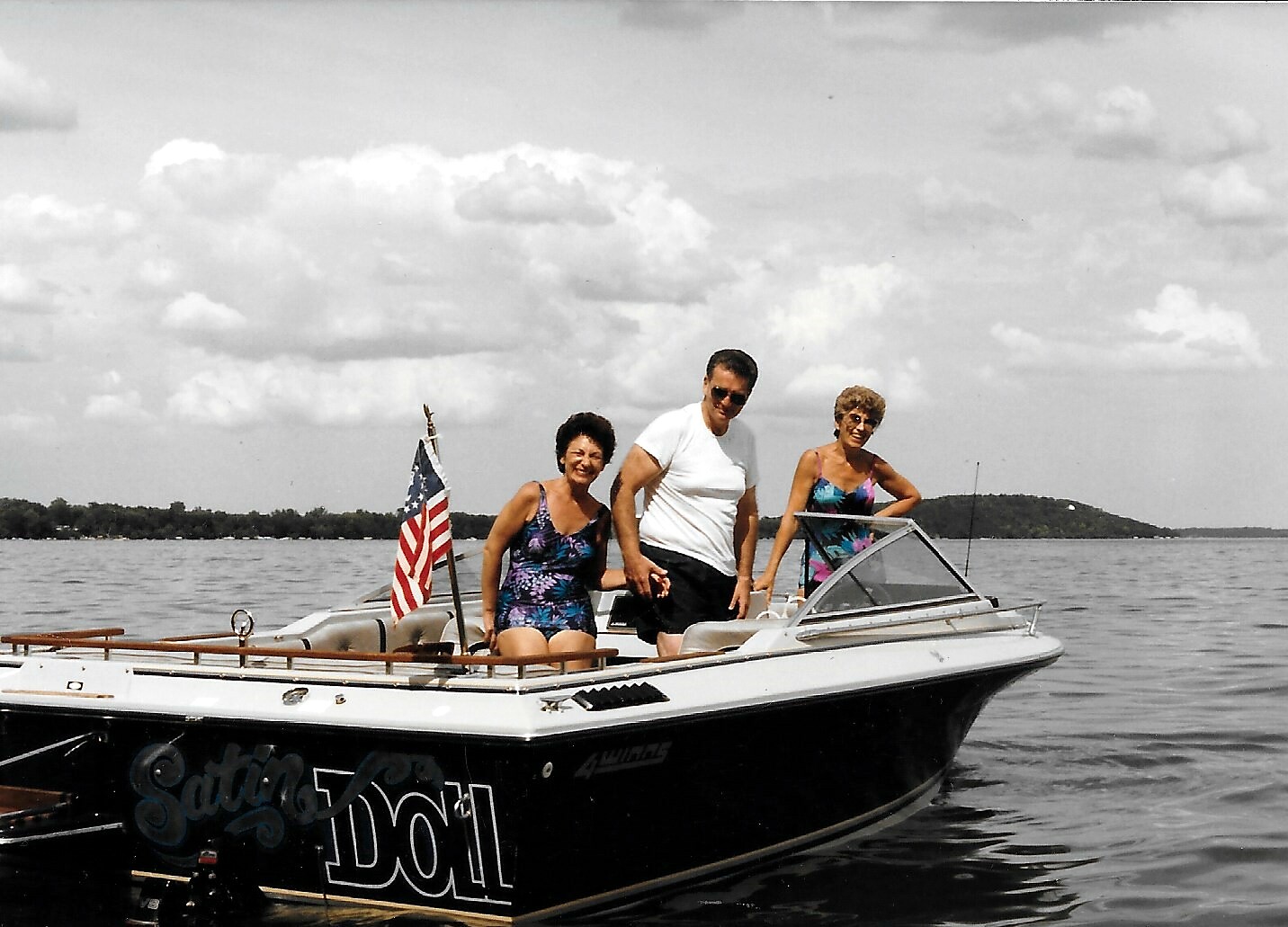 Three people in swimsuits stand on a white speedboat on a calm lake, an American flag flying at the stern under a cloudy sky.