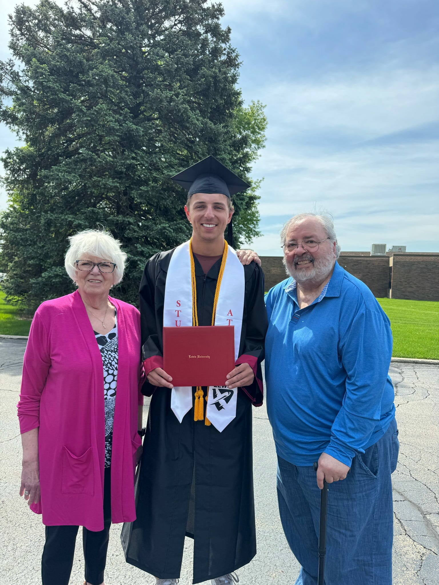 Graduate in cap and gown holding diploma, flanked by a smiling woman in pink and a man in blue outdoors under a blue sky.
