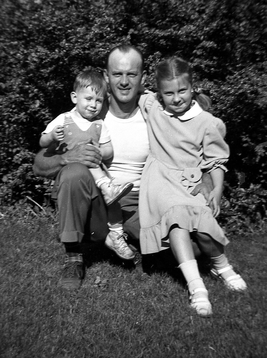 Father sits on the grass with his two children, a boy on his lap and a girl beside him, outdoors in a garden/yard setting.