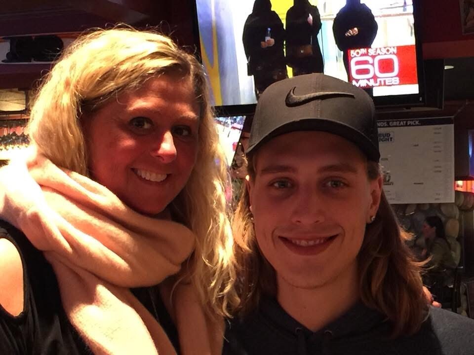 Two smiling people posing indoors: a blonde woman with a pink scarf on the left and a man wearing a black Nike cap on the right