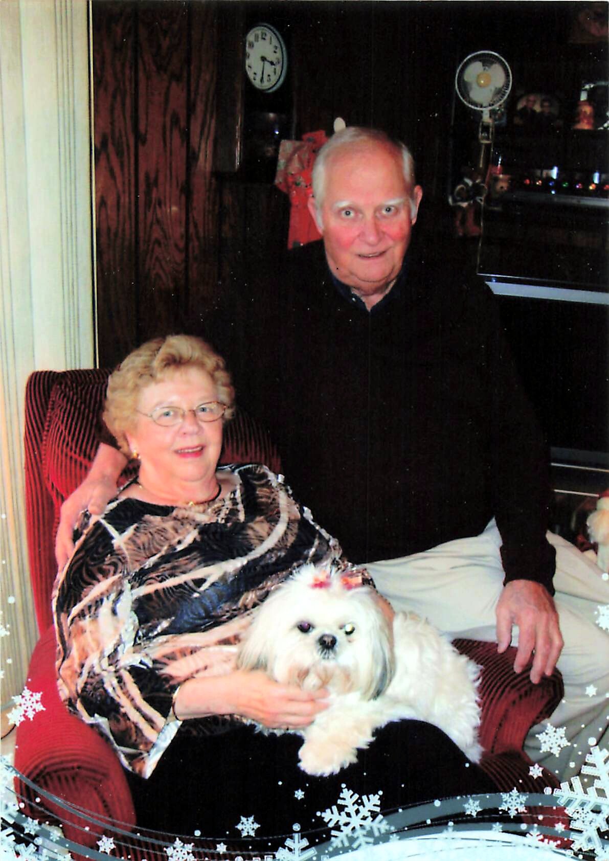 An elderly couple sits together on a red striped chair with a small white fluffy dog on the woman's lap; photo has festive snowflake decorations in the corner.