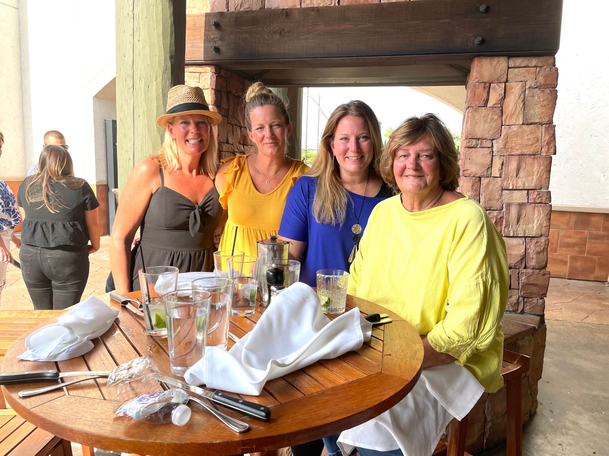 Four women posing together at an outdoor restaurant table with drinks and napkins on the table.