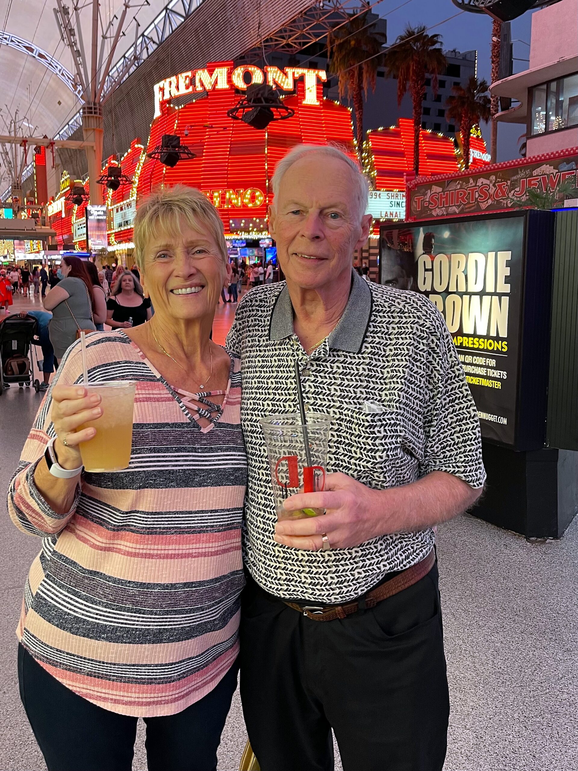 Smiling couple poses with drinks in hand on a neon-lit Las Vegas street, Fremont Street sign behind them.