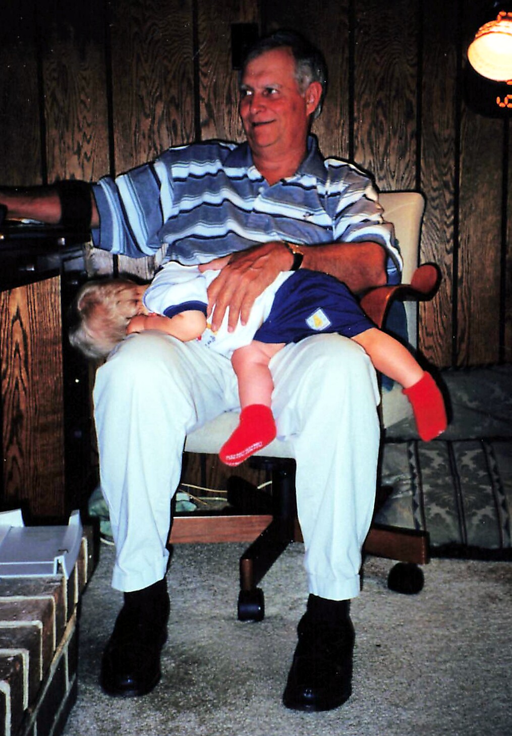 Older man in a striped polo sits in a chair with a small child resting on his lap; red socks on the child.