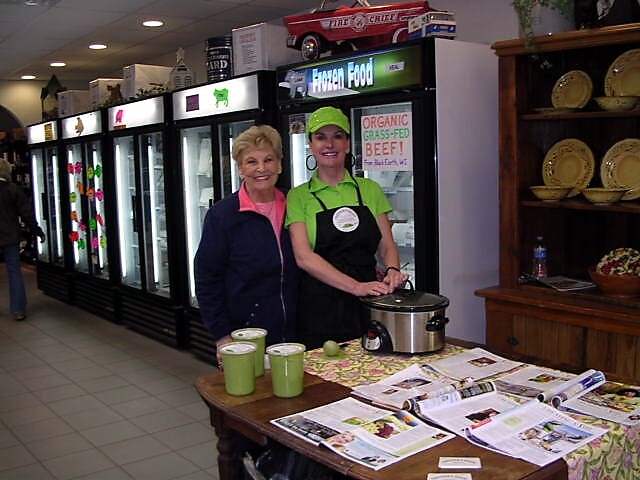 Two women stand at a promotional table with a slow cooker in a grocery store, refrigerators behind them and signs advertising organic beef in the background.