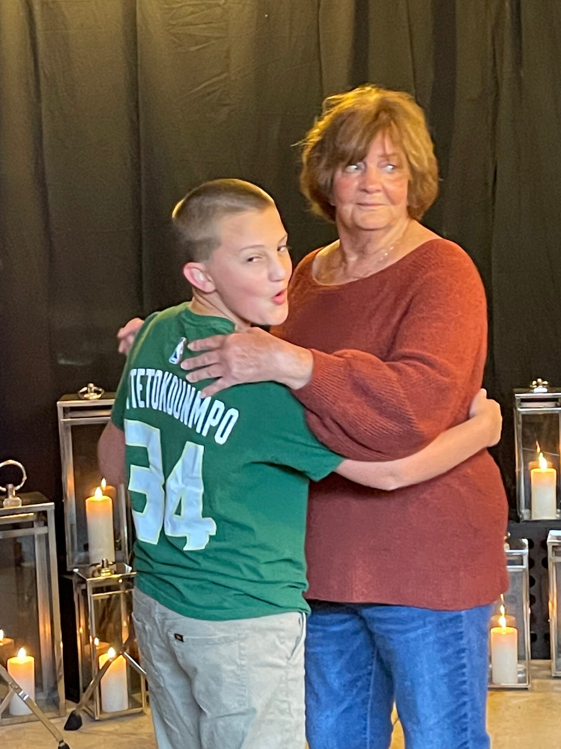 Boy in a green shirt hugs an older woman in a red sweater, both smiling, beside candle-lit glass lanterns.
