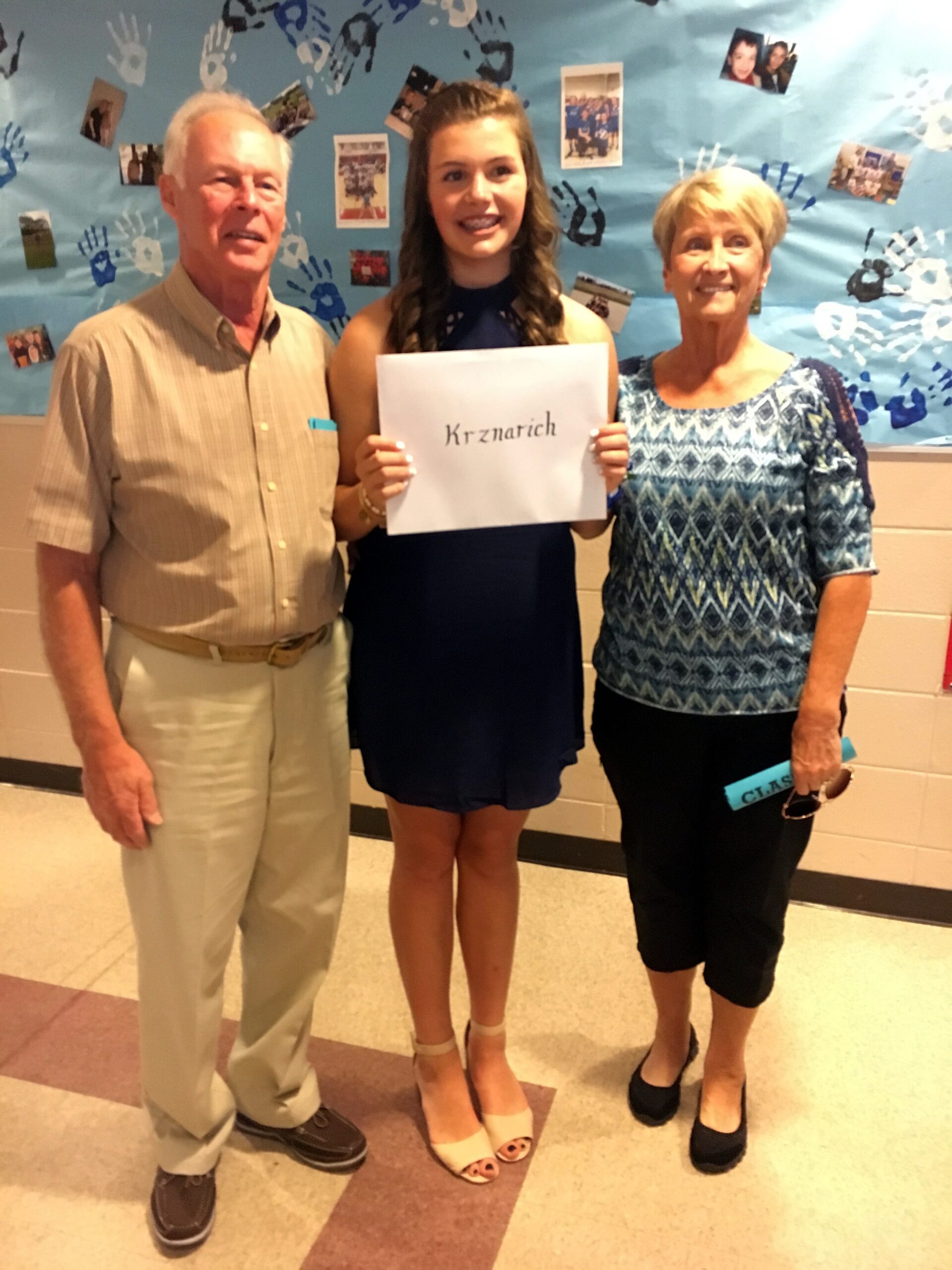 Three people posing indoors: an older man on the left, a teenage girl in the center holding a sign with the word 'Krznarich', and an older woman on the right.