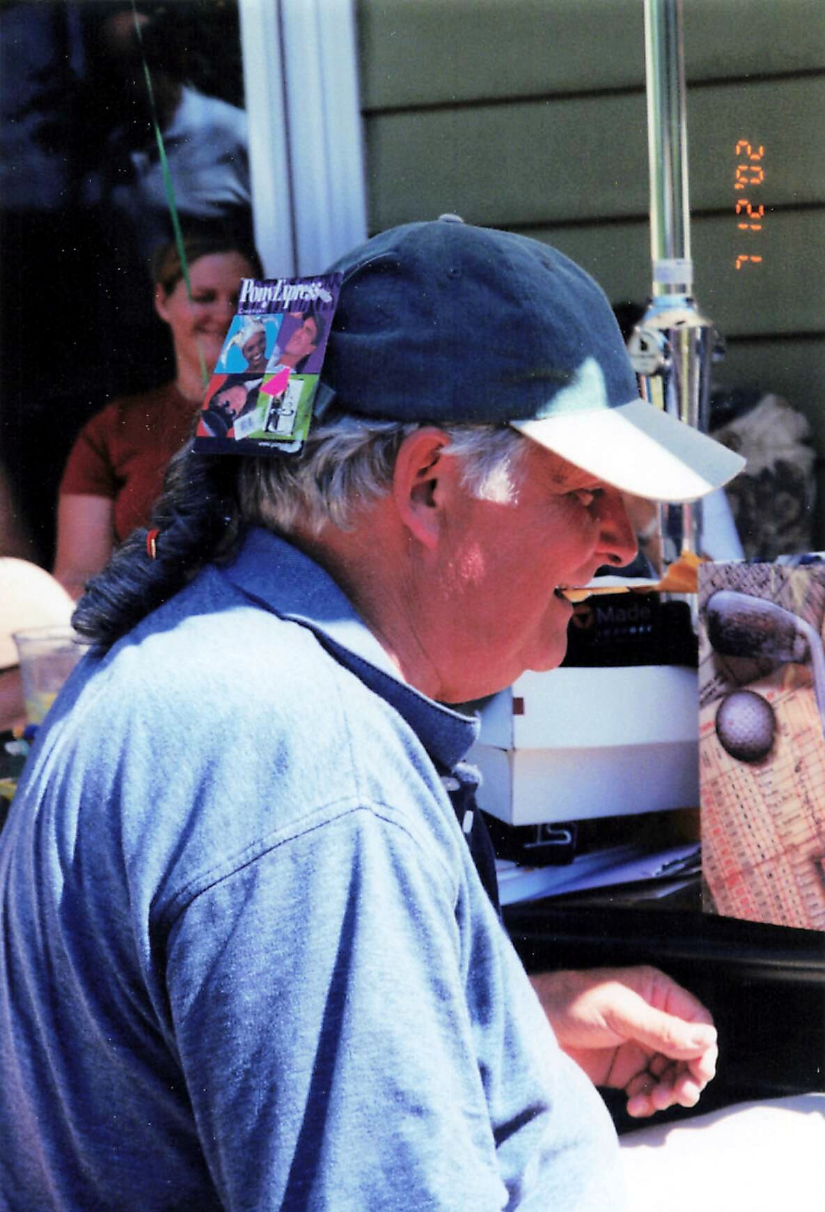 Older man in a blue shirt wearing a dark baseball cap with a small magazine tucked behind the ear, photographed in profile at an outdoor market stall.