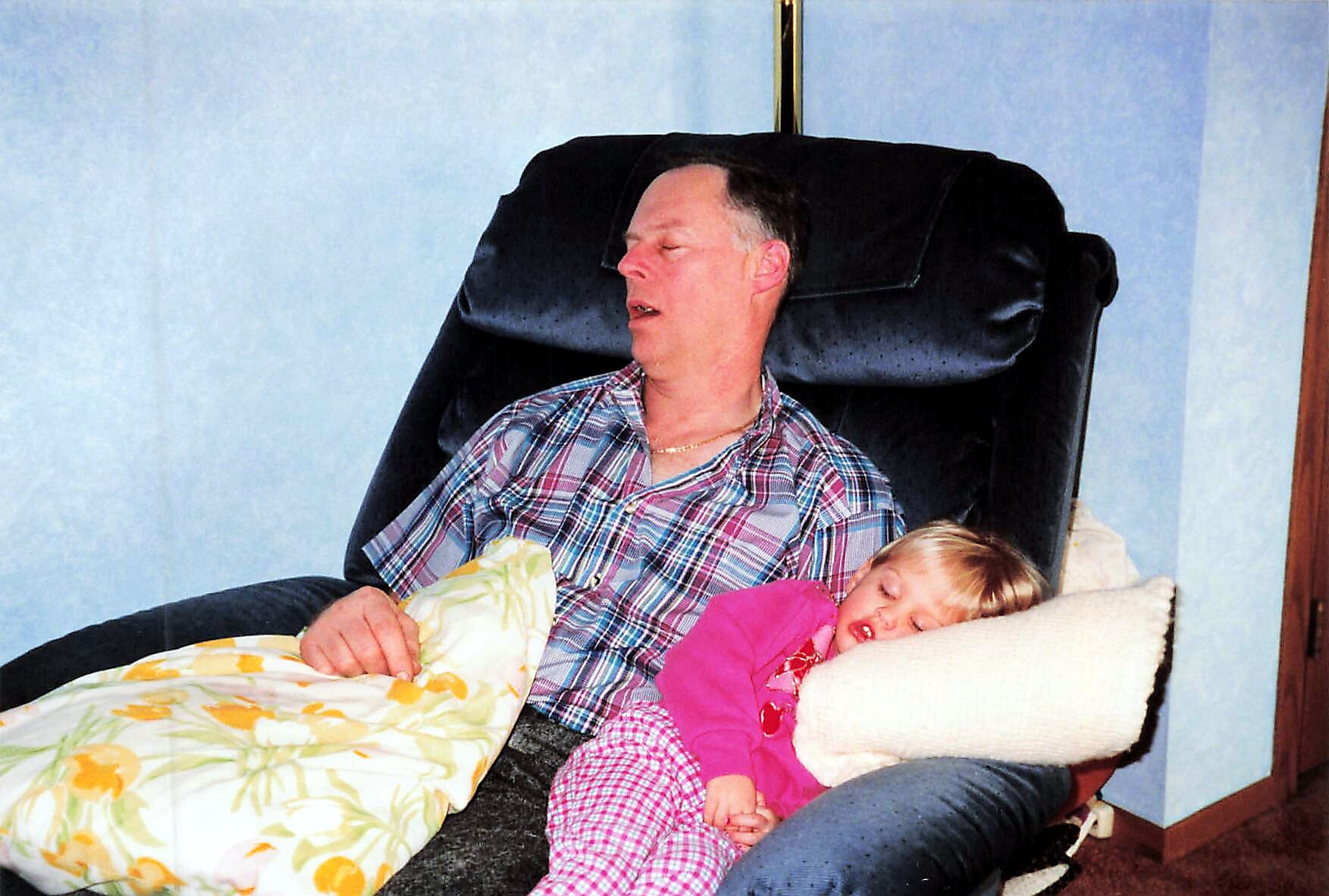 Man dozing in a dark recliner with a child asleep on his lap, both covered by a floral blanket in a living room.