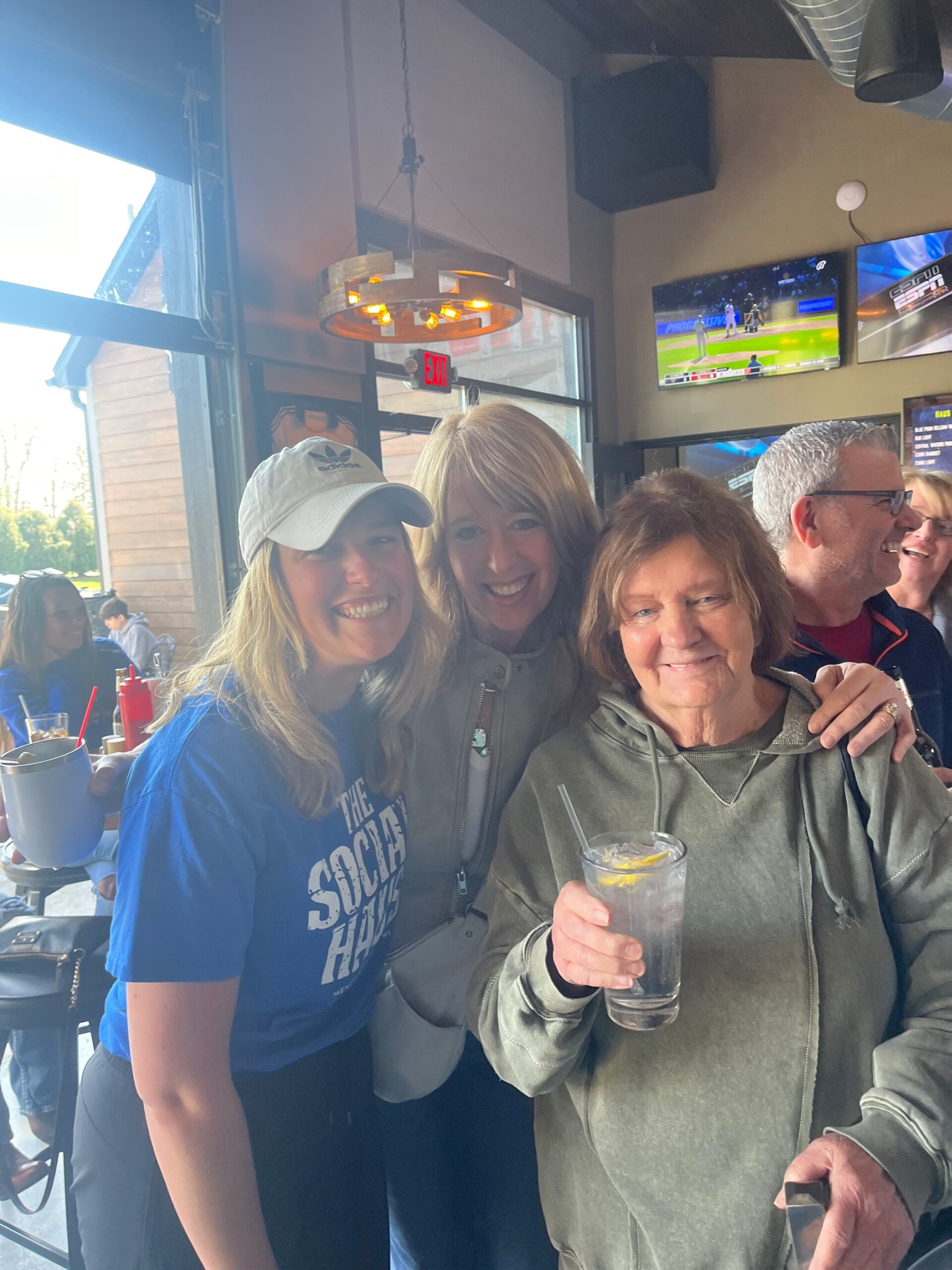 Three smiling people posing for a photo inside a casual bar, drinks in hand, TV screens visible in the background.