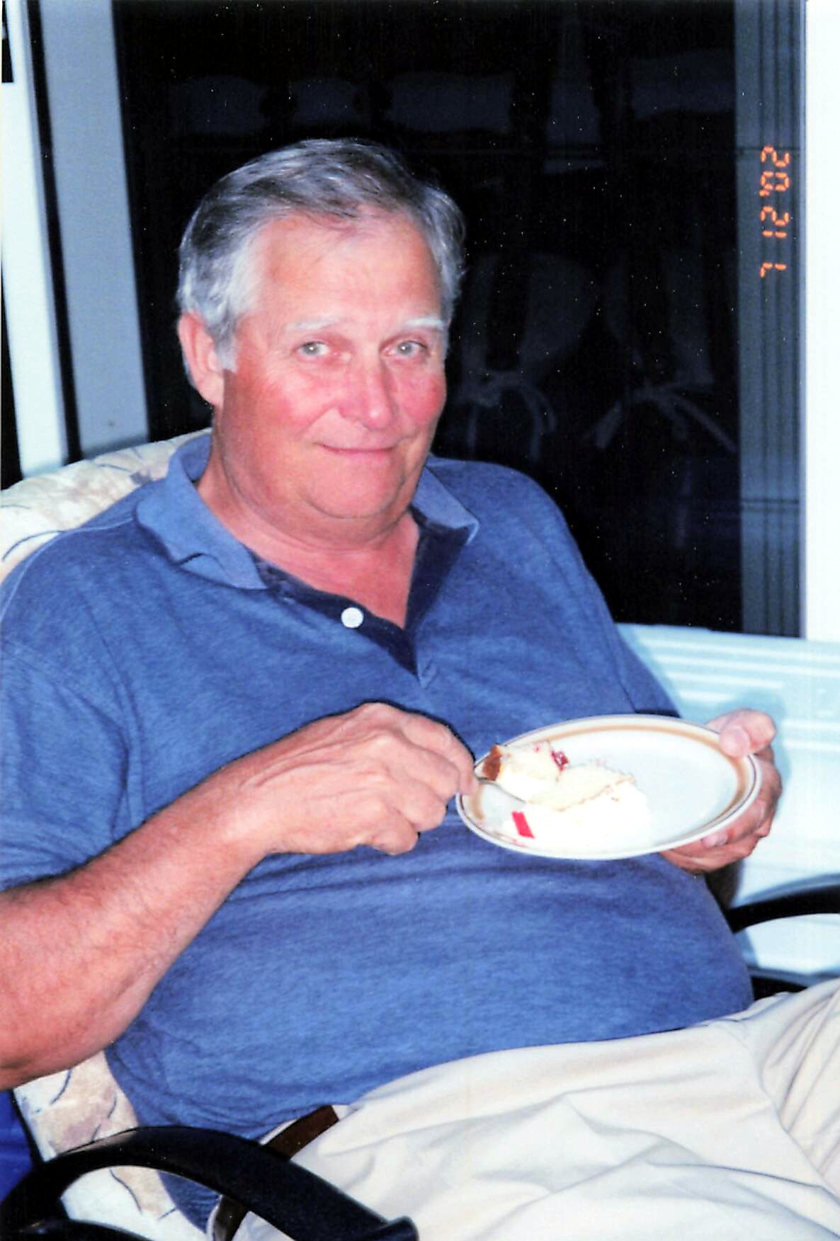Smiling elderly man in a blue polo shirt sits in a chair and holds a plate with a slice of dessert, about to eat with a spoon on his lap.