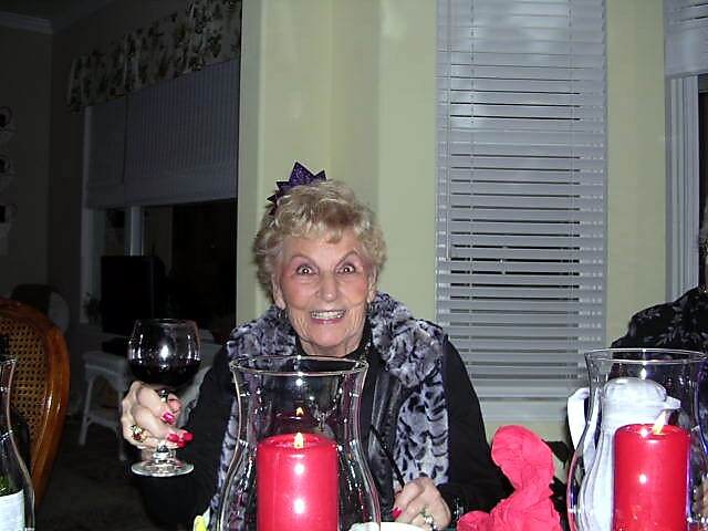 Smiling elderly woman at a dinner table raises a glass of red wine, with lit red candles in a glass hurricane nearby.