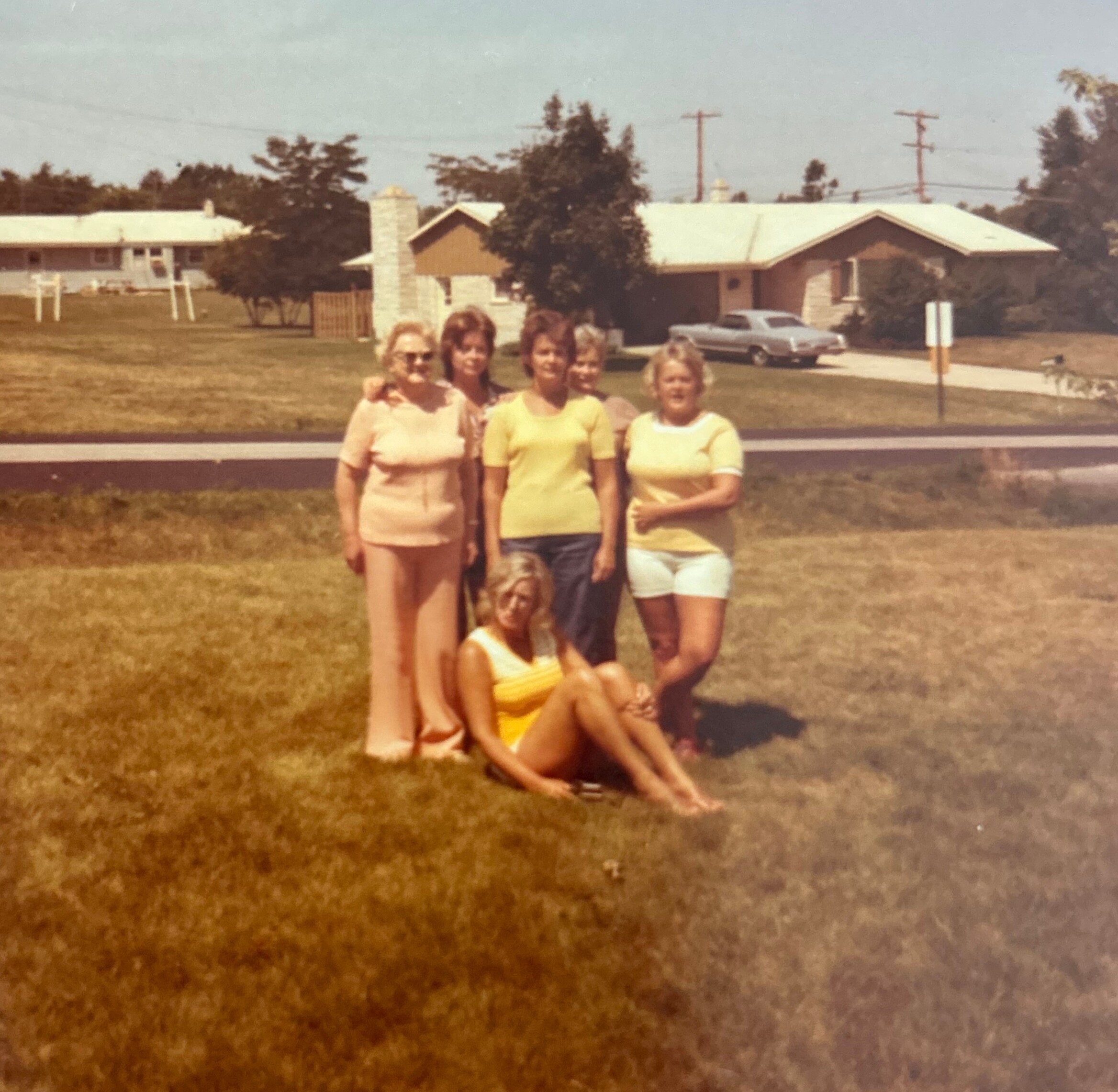 Group of people posing together in a sunny suburban yard, wearing yellow and light clothing, with houses and a car in the background.