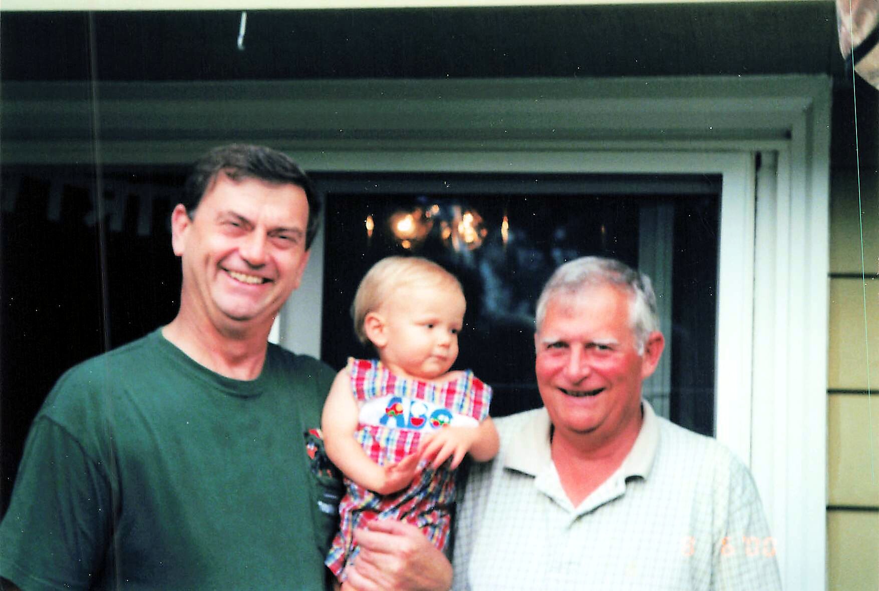 Three people posing in front of a house: a man in a green shirt on the left, a baby girl in a plaid dress held by an older man on the right, all smiling.