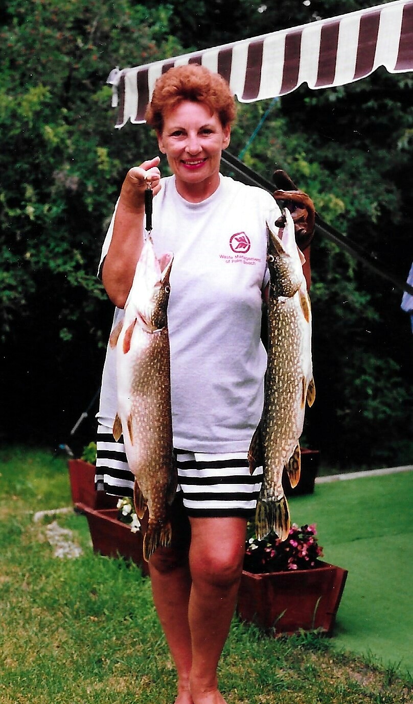 Woman standing on grass outdoors, holding two large fish by the mouths with a striped canopy behind them.