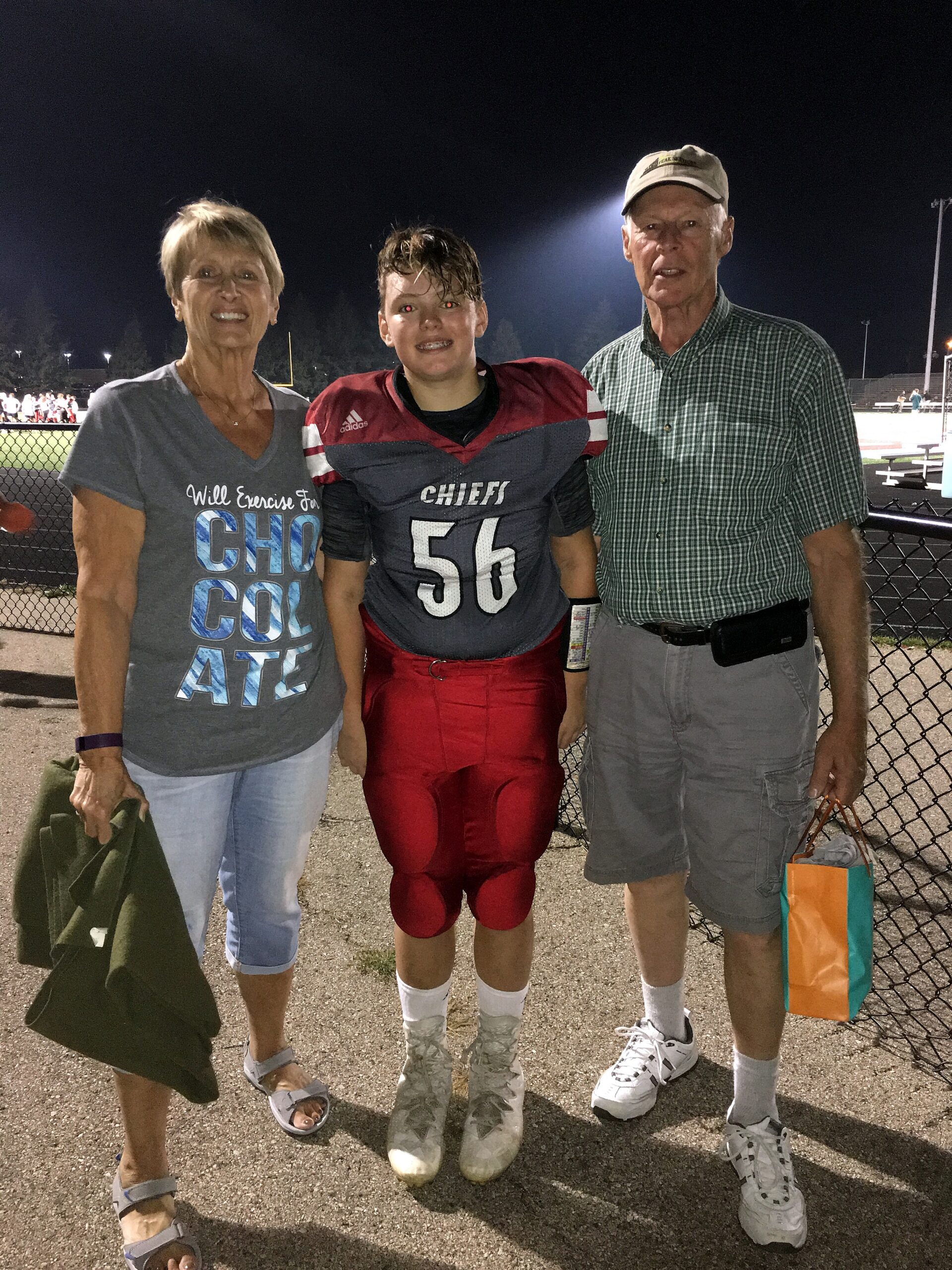 Young football player in a red Chiefs uniform (number 56) stands between smiling woman and man at night near a chain-link fence.