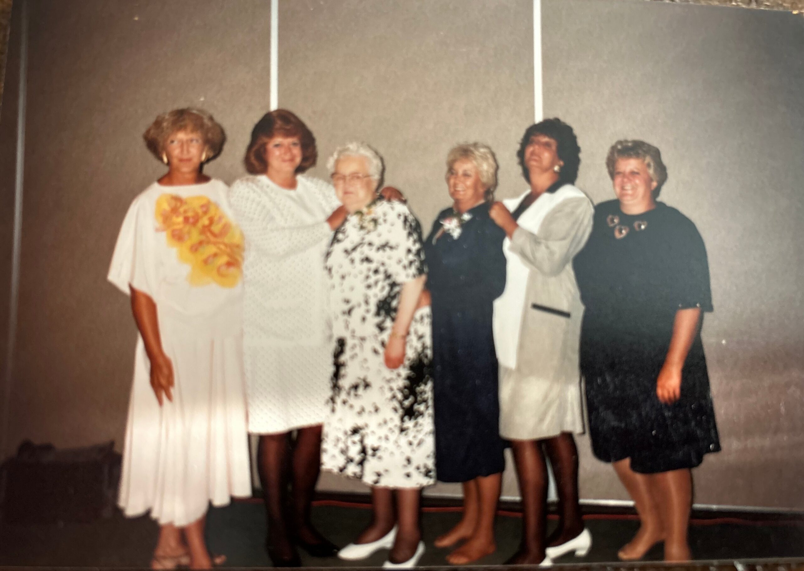 Six women stand together in a row, posing for a group photo in dresses of white, floral, and dark outfits on a simple backdrop.