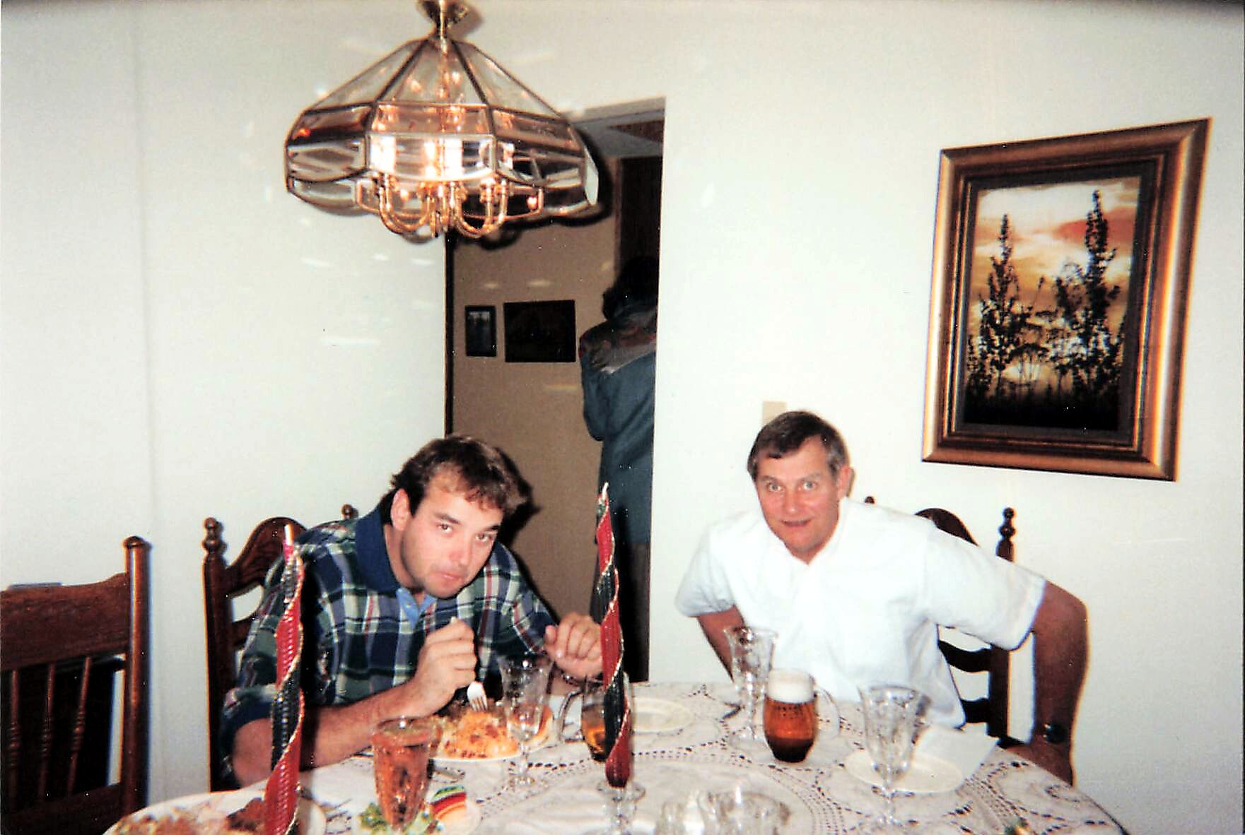 Two men sit at a dining table during a celebration, with candles and party decorations visible.