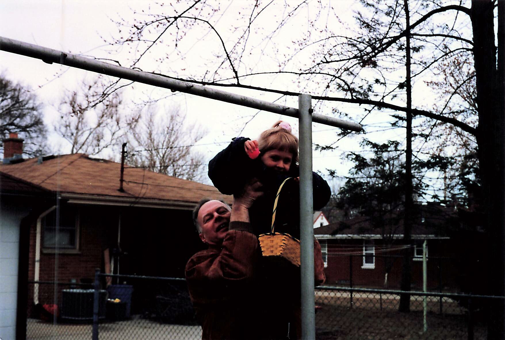 Man lifting a child onto a metal play frame in a backyard, with a woven basket hanging from the pole nearby.