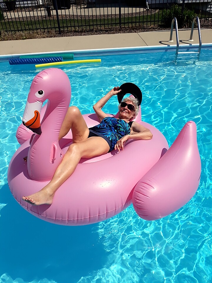 Older woman relaxing on a pink flamingo pool float, wearing sunglasses and a blue swimsuit, in a sunny pool.
