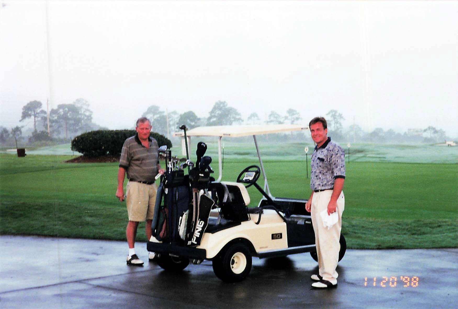 Two men stand beside a golf cart loaded with golf bags on a wet golf course, with trees in the distance.