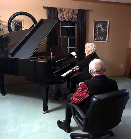 Two elderly people at a black grand piano in a living room; one plays the piano while the other sits in a chair nearby.