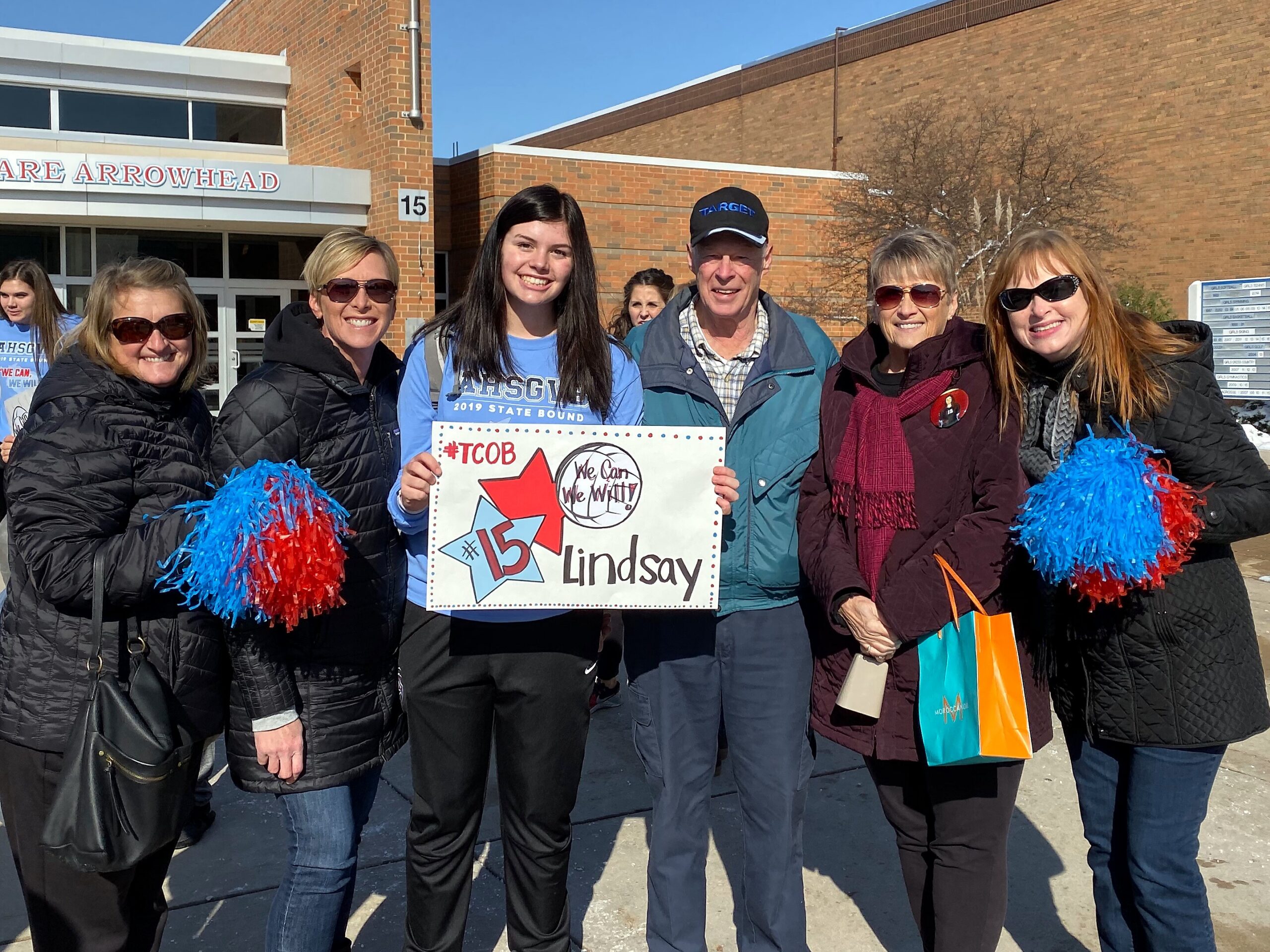 Group of adults and a teenage girl smiling outside a brick school building, holding a sign that reads '#15 Lindsay' with a red star graphic and a circular logo nearby.