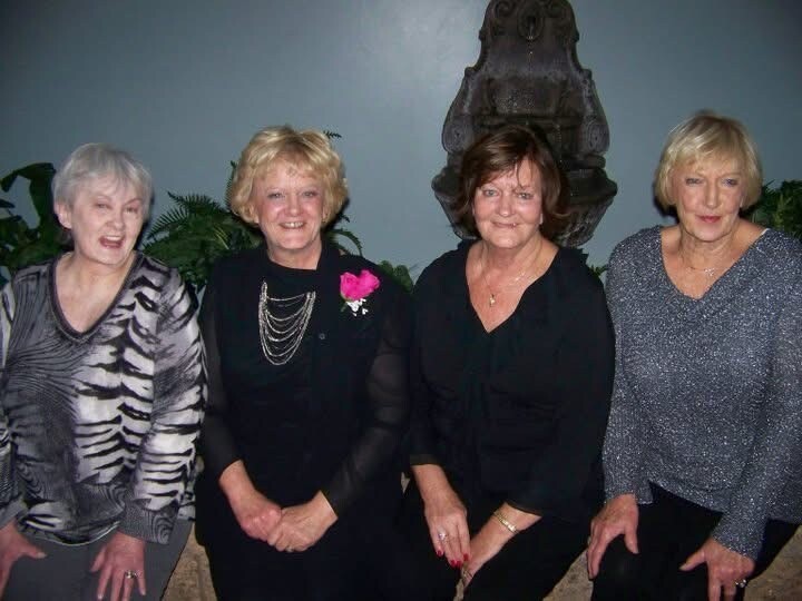 Four smiling older women sit side by side indoors, dressed up for a gathering; one wears a pink corsage and layered necklace.