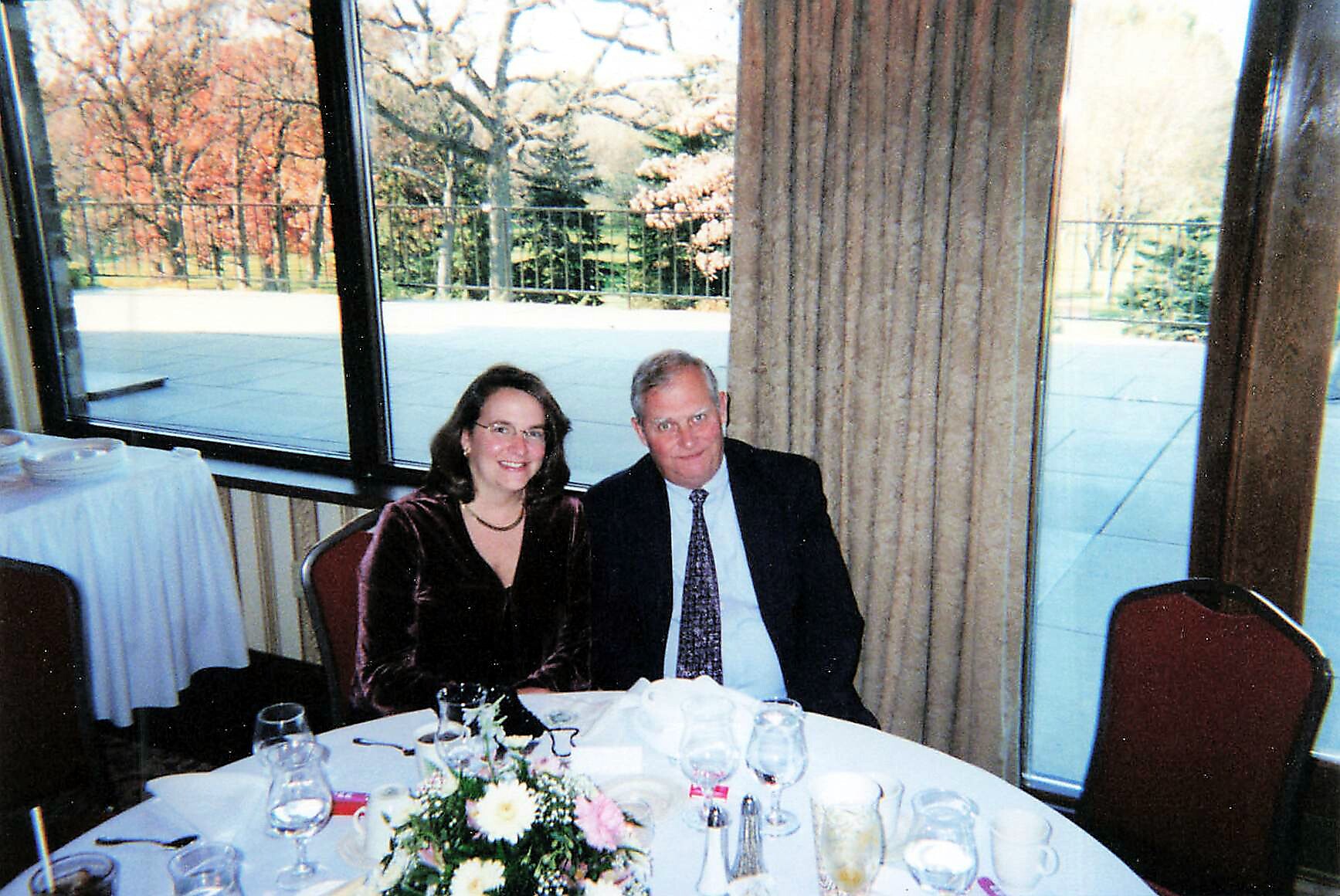 Smiling couple seated at a round banquet table with a floral centerpiece during a formal event by large windows.