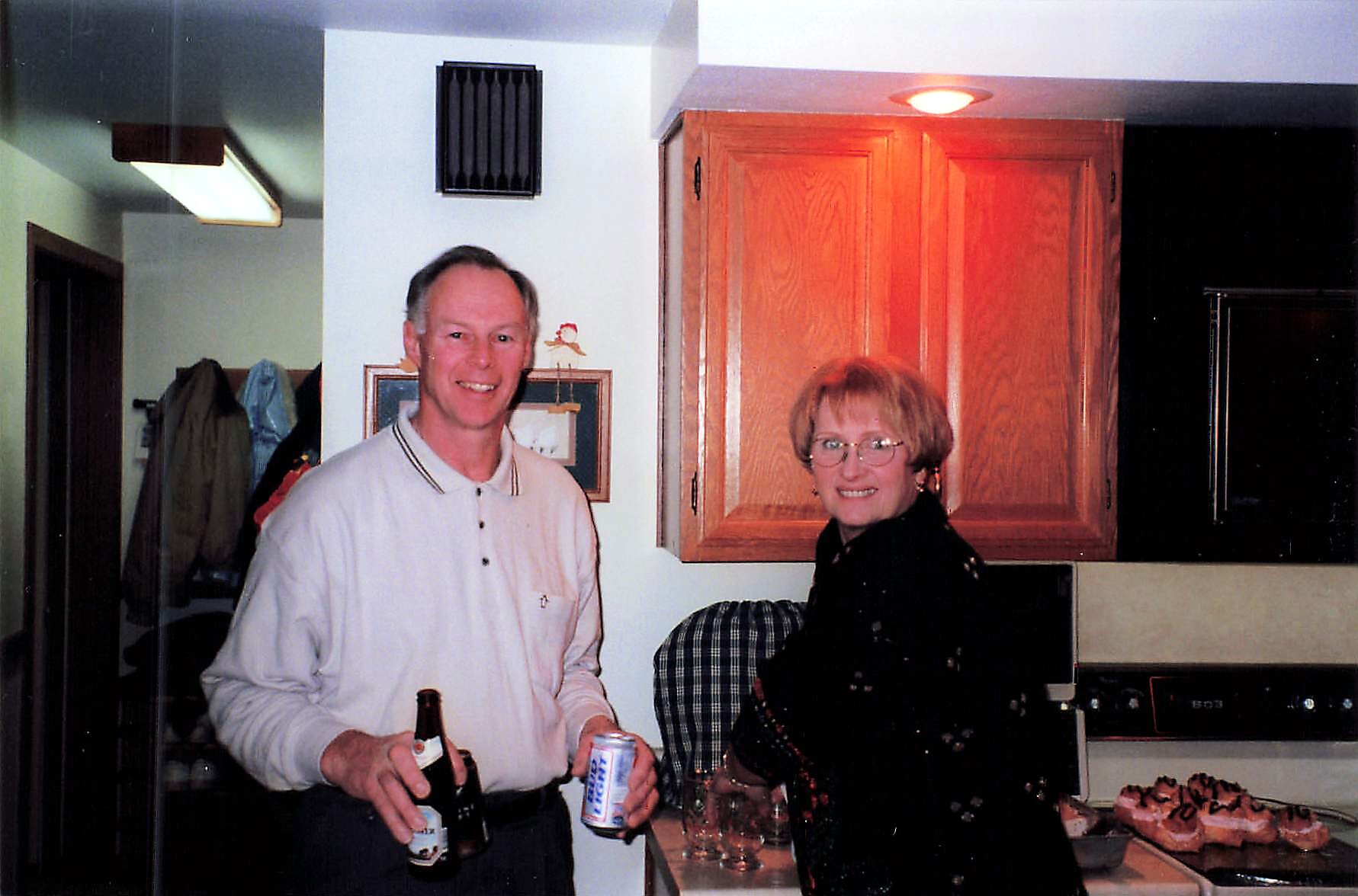 Smiling man in a white polo and a woman with glasses stand in a kitchen, holding drinks with donuts on the counter nearby.