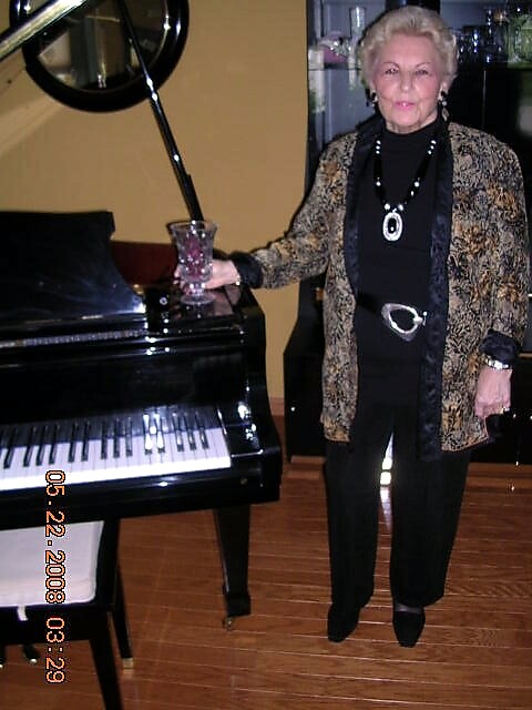Older woman with short blonde hair in a gold-patterned jacket stands by a black grand piano, holding a glass.