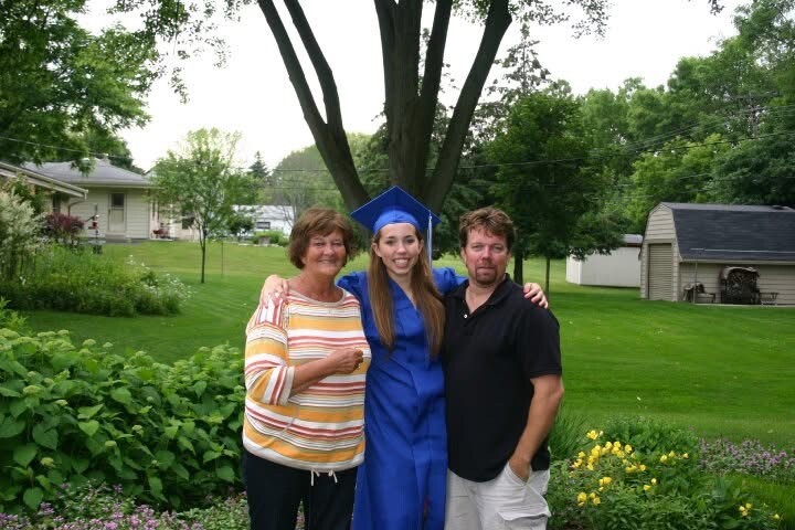Graduate in blue cap and gown posing with her parents in a green suburban yard, smiling at the camera.