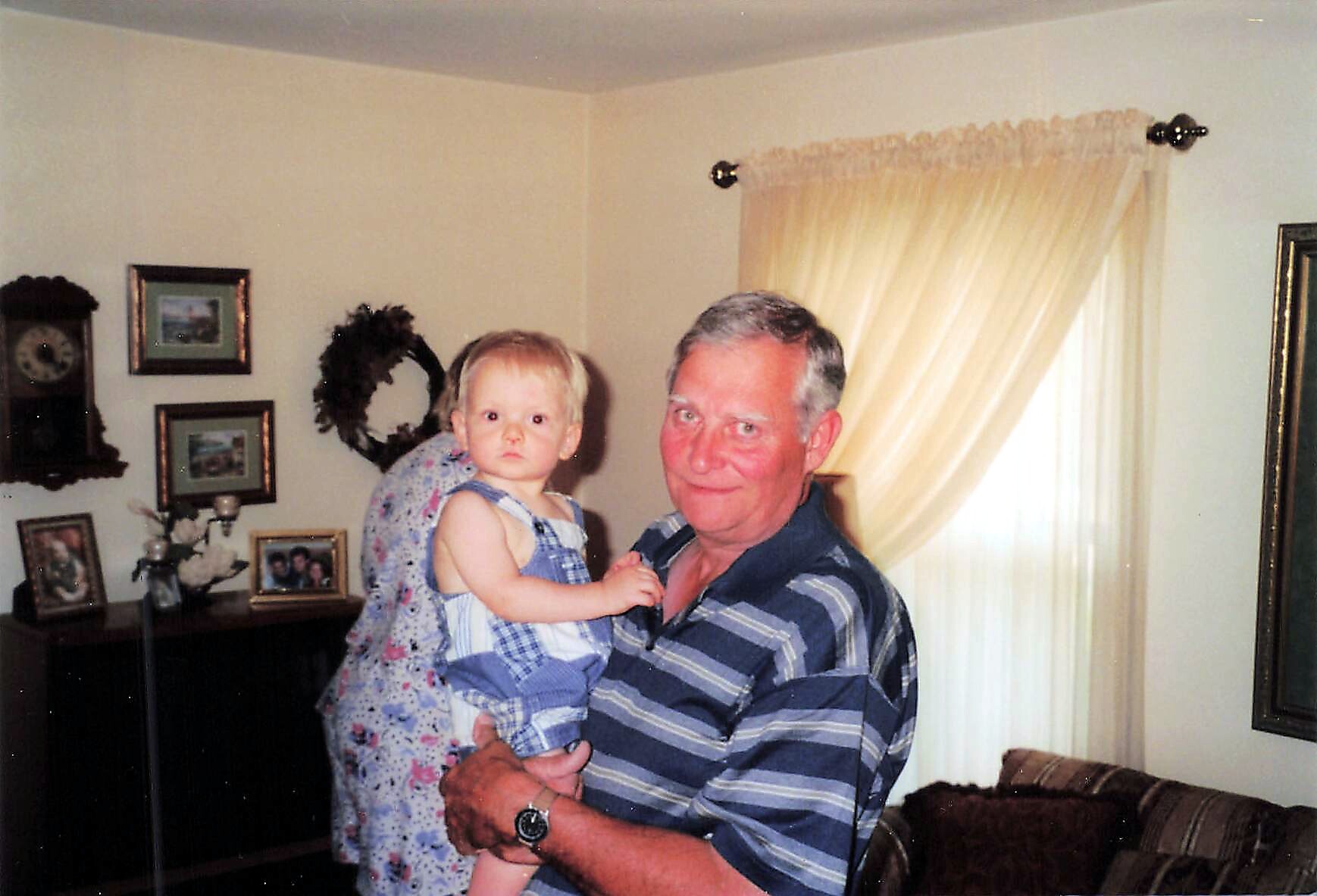 Grandfather holding a young blonde baby girl in a cozy living room with family photos on the wall behind them.