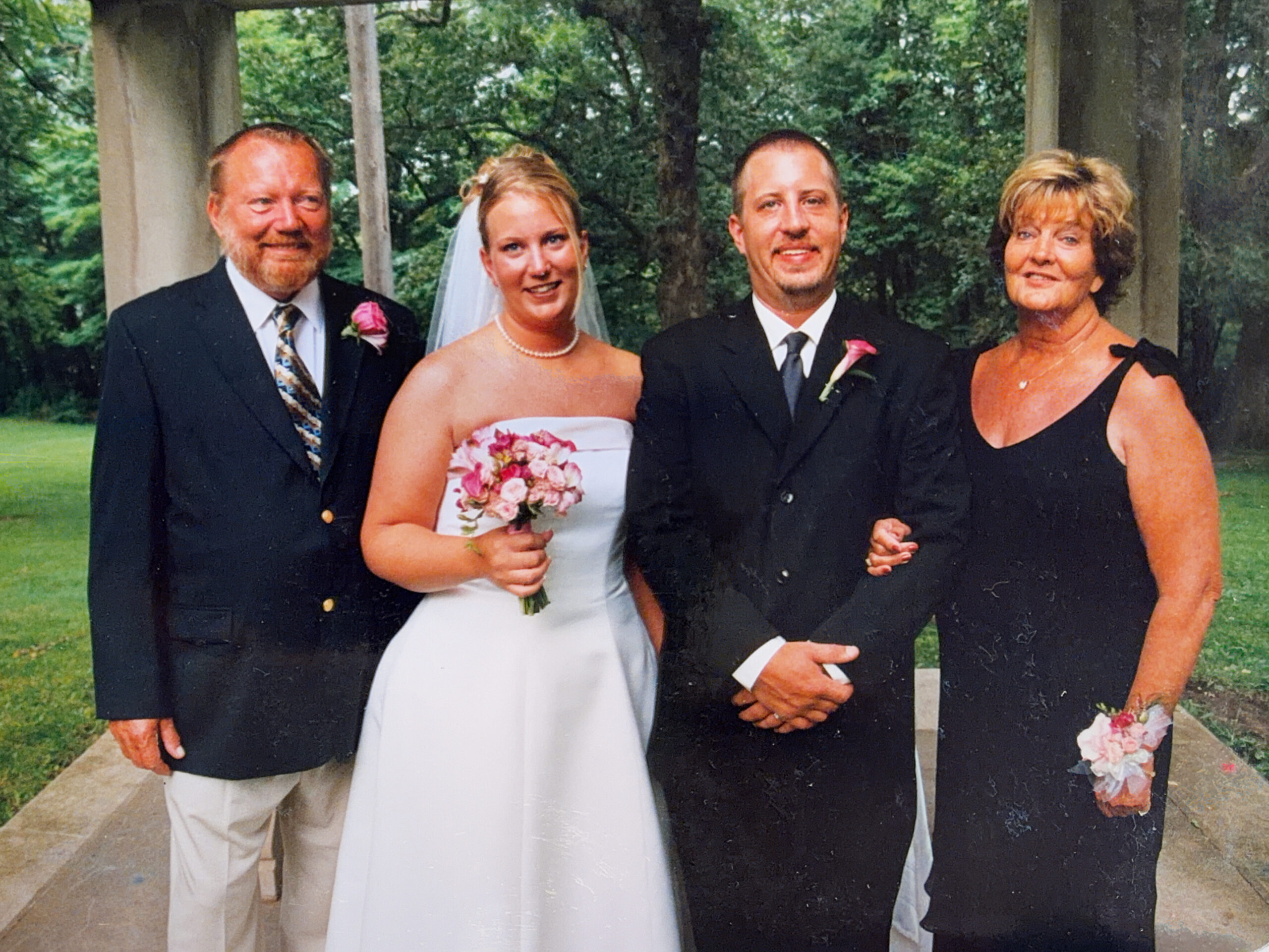 Wedding party portrait outdoors under a gazebo: bride in a white strapless gown holding a pink bouquet, groom in a black suit with a pink boutonniere, and two older adults in formal attire on either side.
