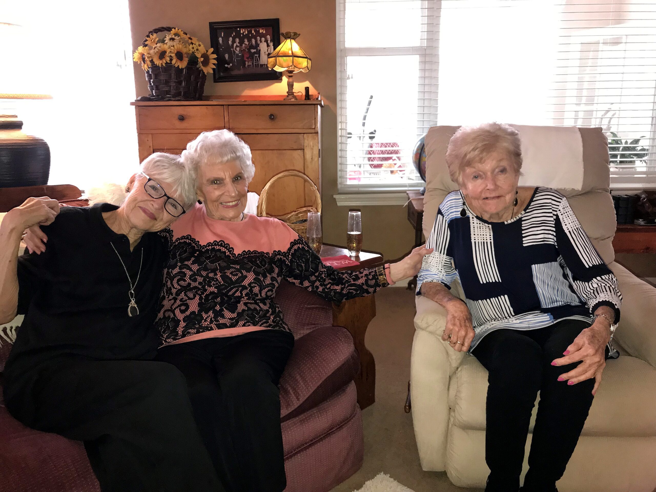 Three elderly women sit together in a living room, smiling at the camera, with a wooden dresser, sunflower basket, and lamp behind them.