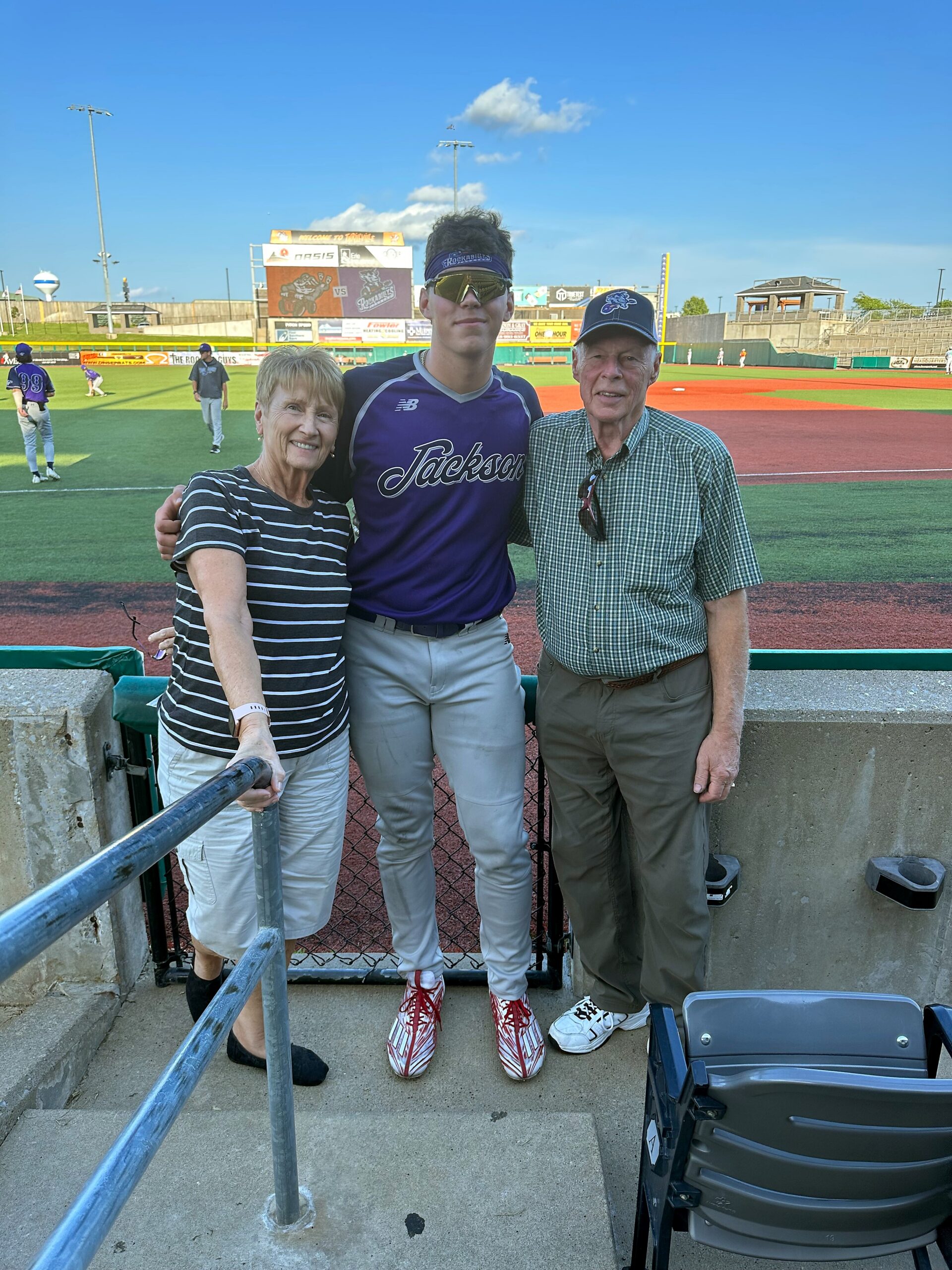 Young baseball player in a purple Jackson jersey stands with an older couple on a stadium railing, posing for a photo with the field behind them.