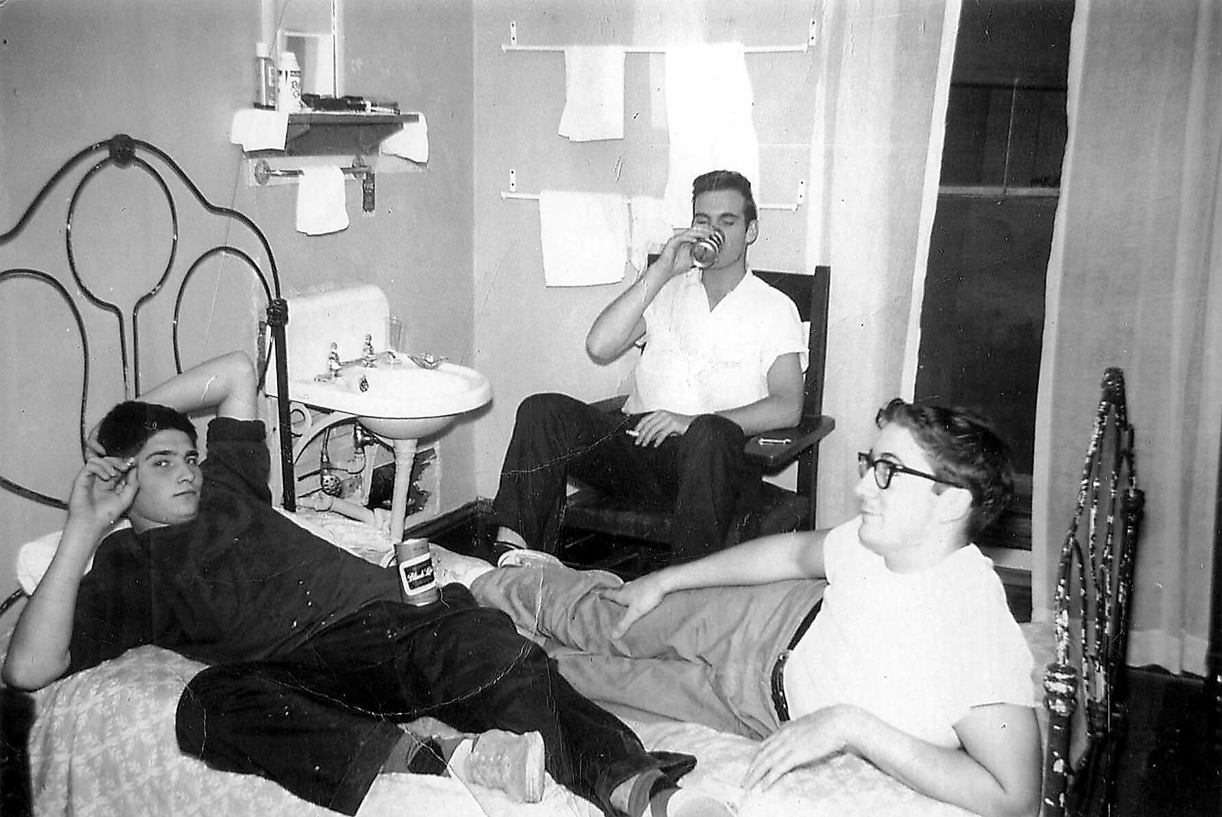 Three young men in a small dorm room: one lying on a bed and drinking, another seated in a chair drinking from a can, and a third smiling in a white shirt near the foot of the bed; a sink and towel rack visible in the background.