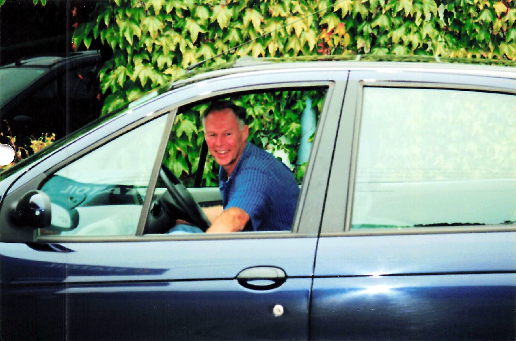 Smiling man in a blue shirt sits in the driver’s seat of a grey car, with a leafy hedge behind the vehicle.