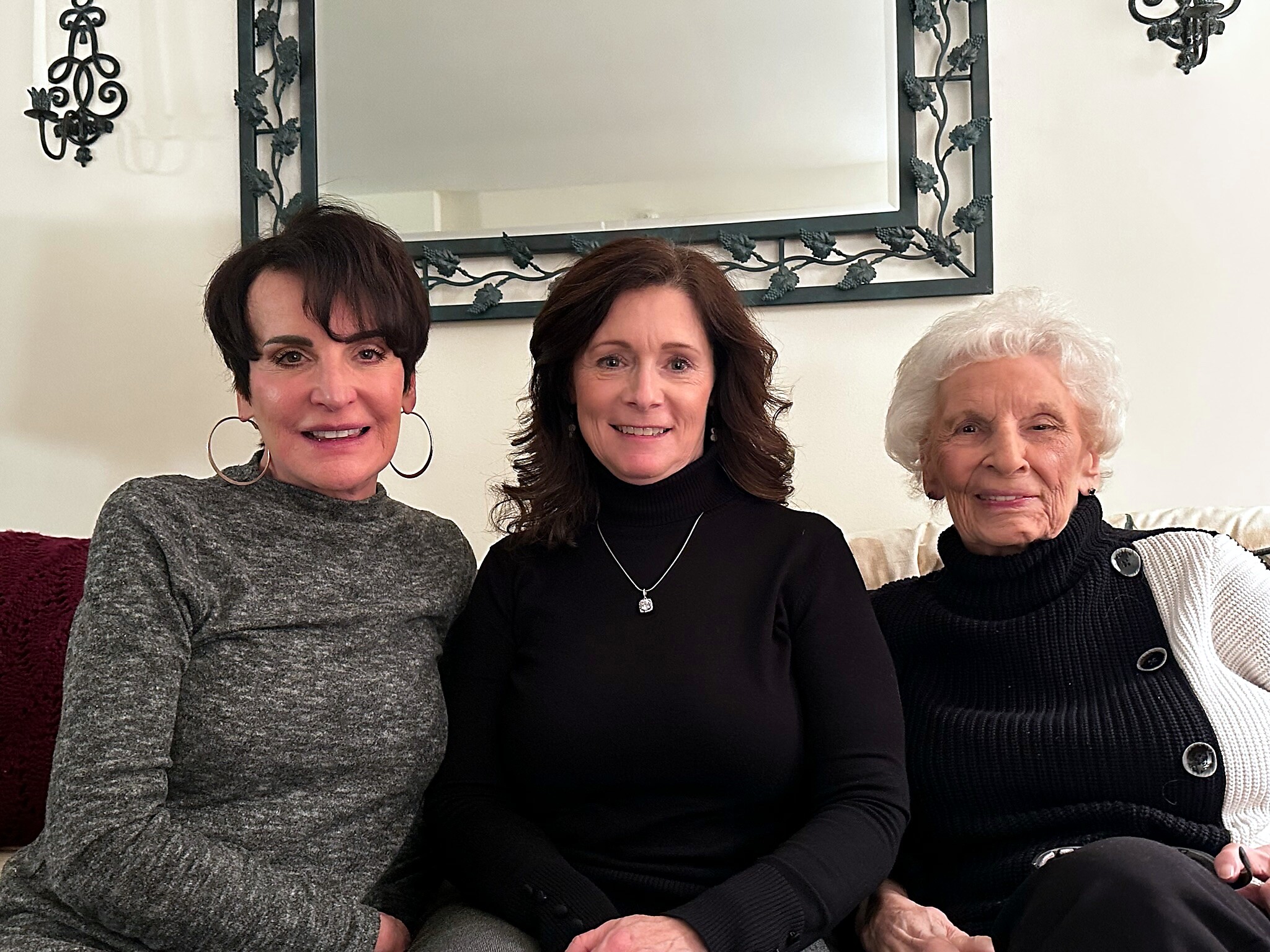Three women sit closely together on a sofa indoors, smiling, with a decorative mirror on the wall behind them.