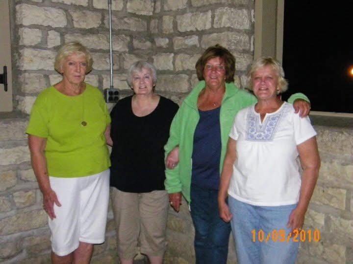 Four older women standing side by side against a stone wall, smiling at the camera.