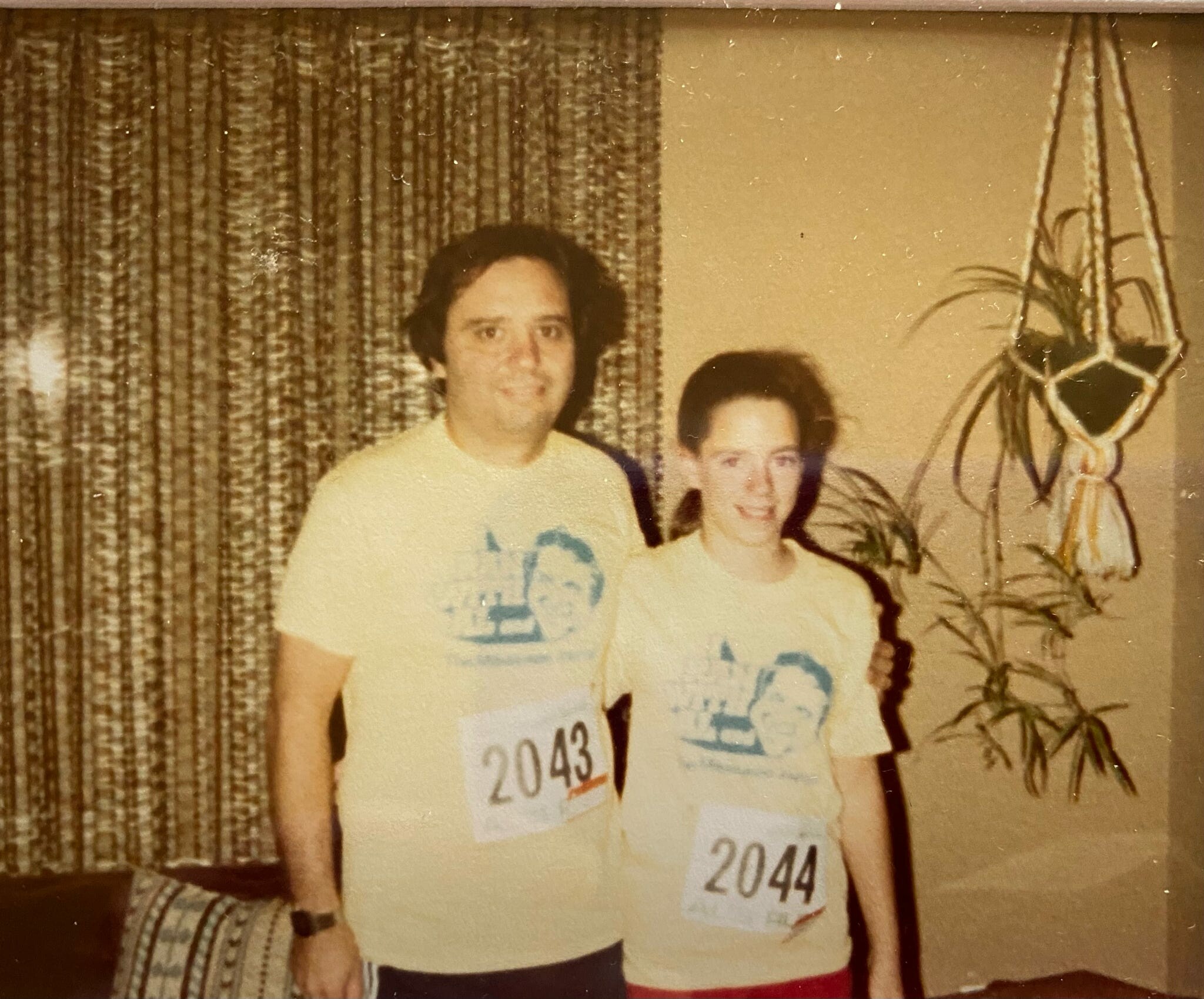 Two people wearing race bibs 2043 and 2044 pose together indoors in front of a patterned curtain.
