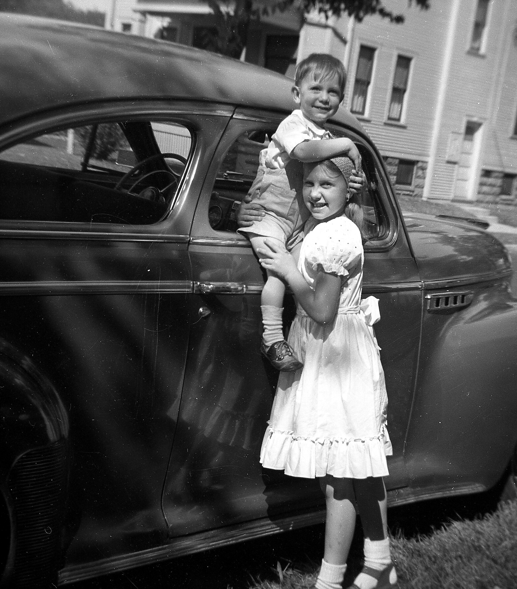 Two children on a vintage car: a smiling toddler sits on the car door while a girl in a dress supports him, in a suburban street setting.