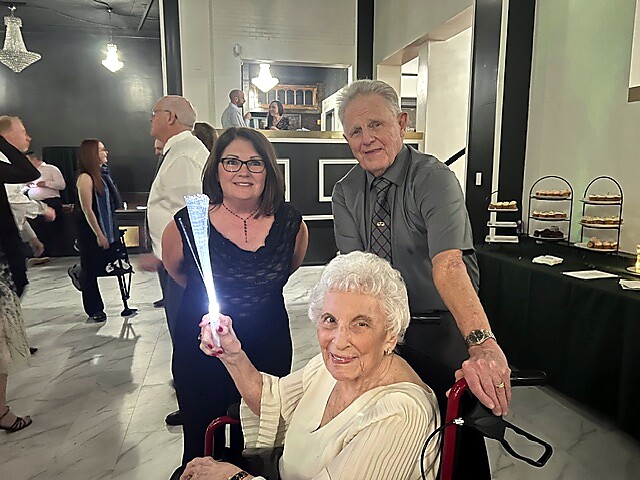 Elderly woman in a wheelchair holds a bright glowing wand as she smiles at a social event with two guests beside her; a cake/coffee table and attendees in the background.