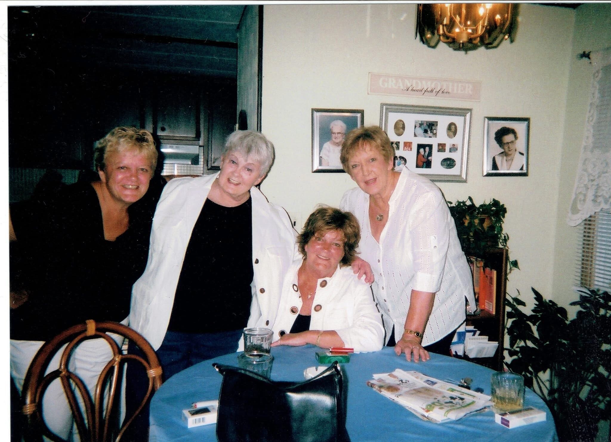 Four women gathered around a blue table, smiling at the camera in a home setting.