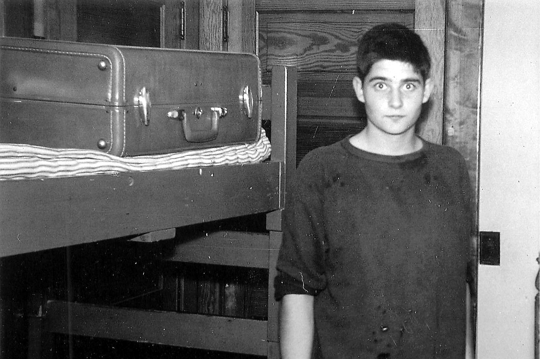 Young man in a dark shirt stands beside a wooden bunk bed with a vintage suitcase on the top shelf.