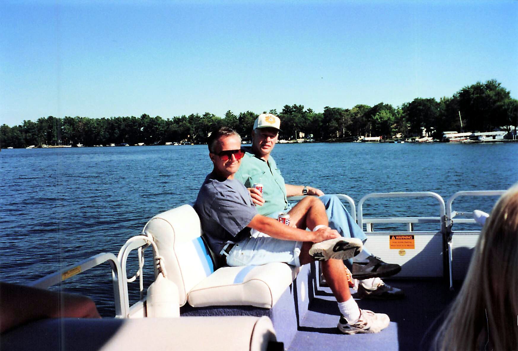 Two men sitting on a pontoon boat on a lake, one wearing sunglasses and a gray shirt, the other in a green shirt and cap, holding drinks.