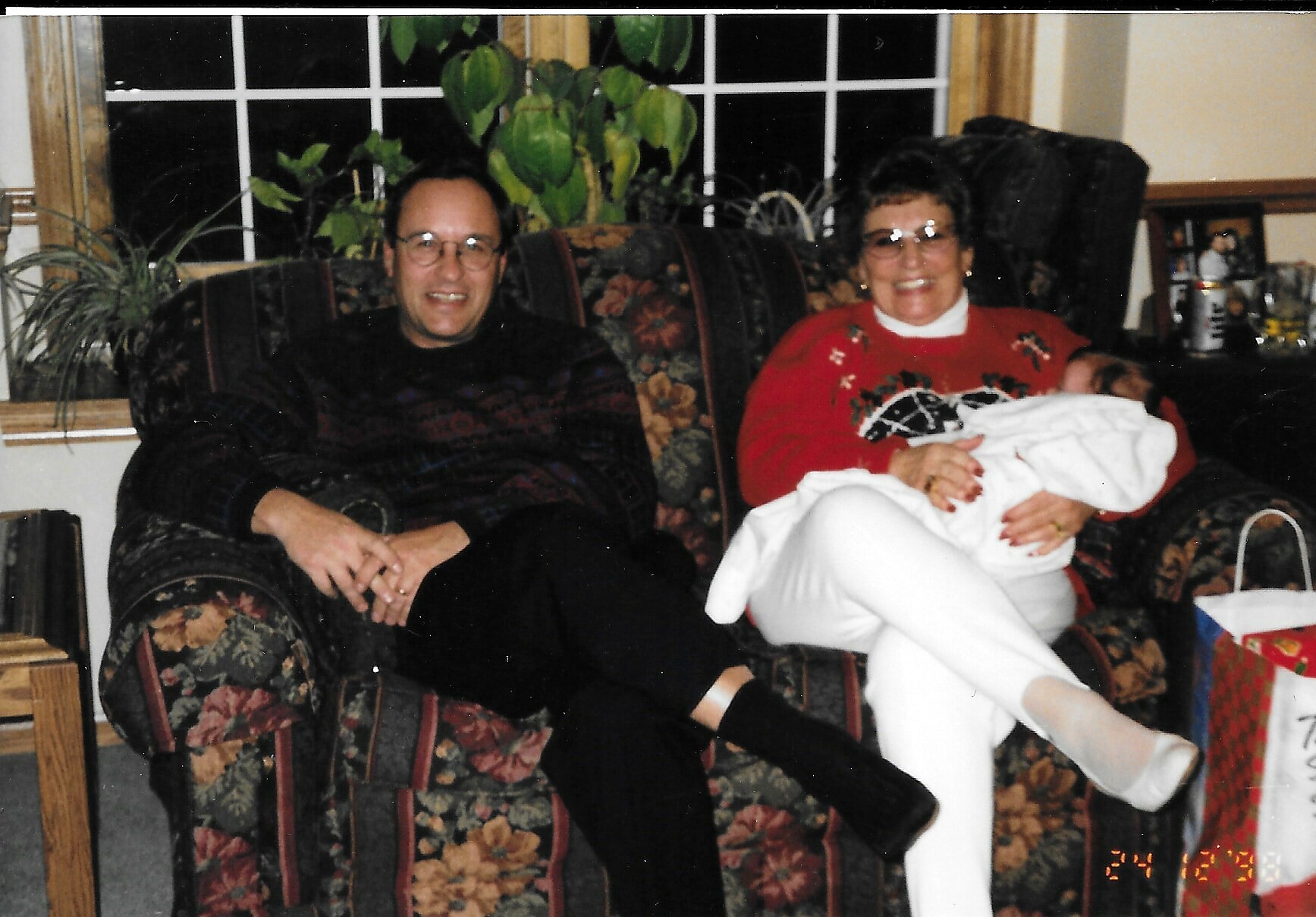 A couple sits on a floral-patterned sofa in a living room, smiling at the camera while the woman holds a newborn wrapped in a white blanket.