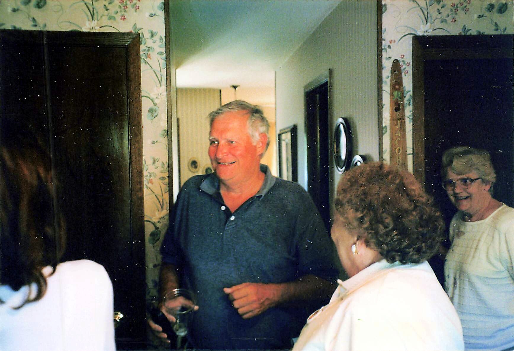 Smiling man in a blue polo converses with guests at a casual indoor gathering in a home.]?